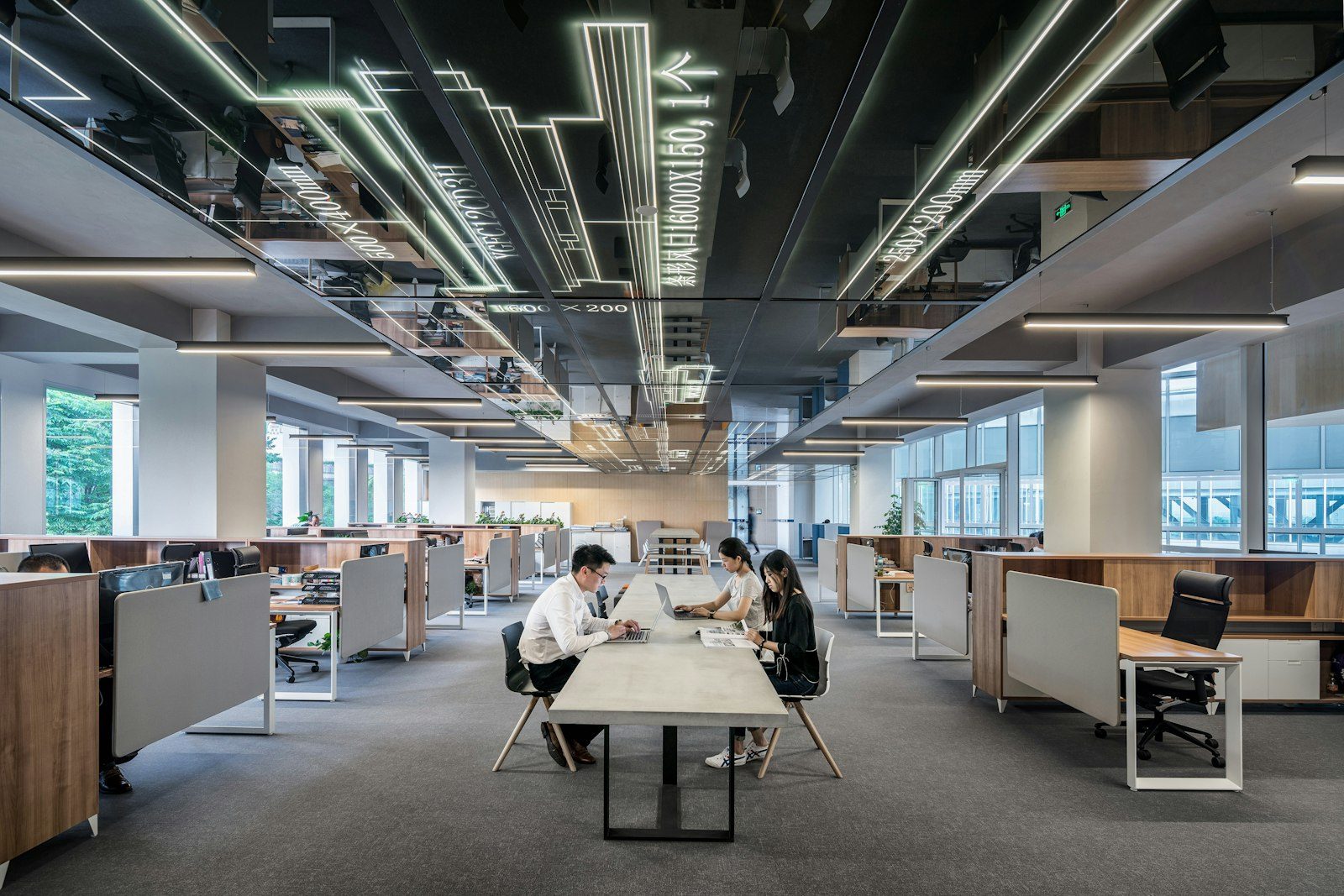 An open-plan office with desk rows along both sides and three colleagues working at a shared central table